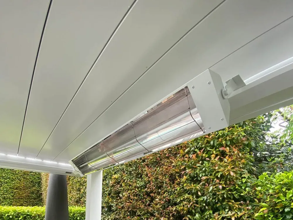 White powdercoated infrared heater fixed under a louvre roof beside a hedge