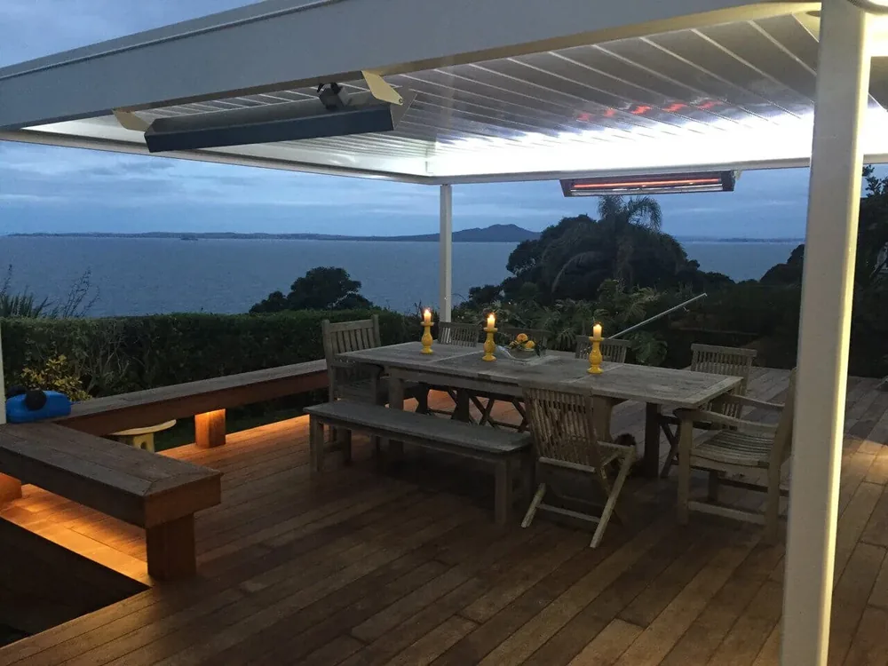 Two heaters under a louvre roof above a deck dining table with a harbour view at dusk
