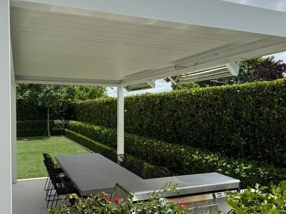 Outdoor heaters under a white louvre roof above an outdoor kitchen and hedged lawn