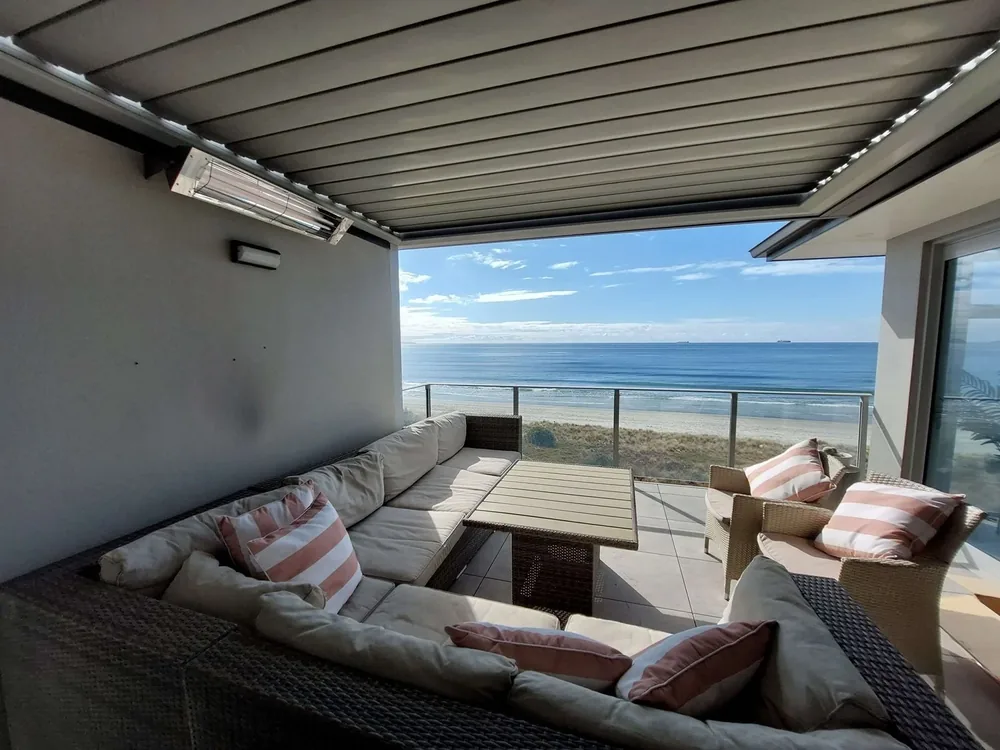 Wall-mounted infrared heater under a louvre roof on a beachfront deck with lounge seating