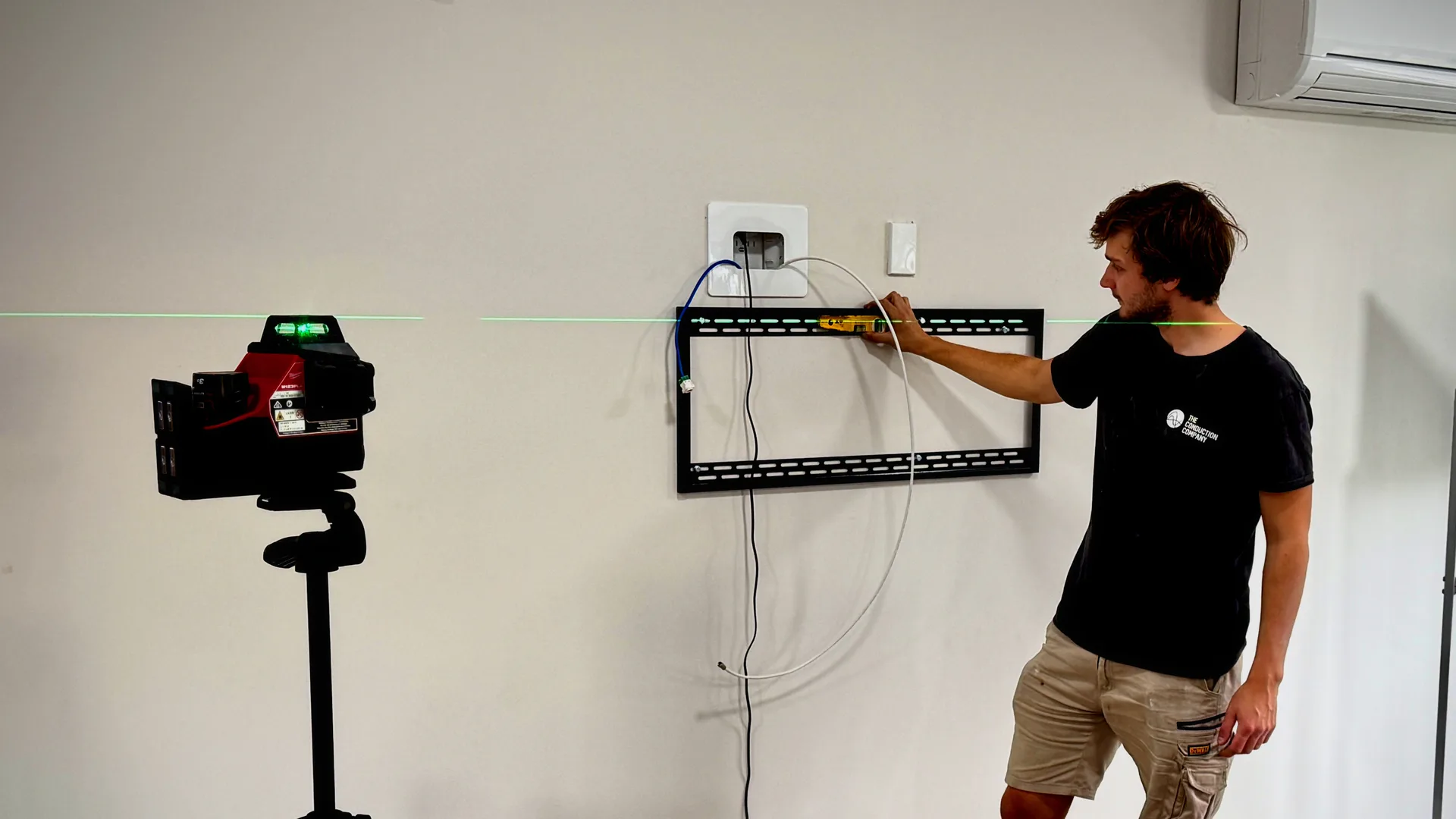 An electrician holding a TV wall bracket to a laser line, with power and data cables run out of a recessed wall box