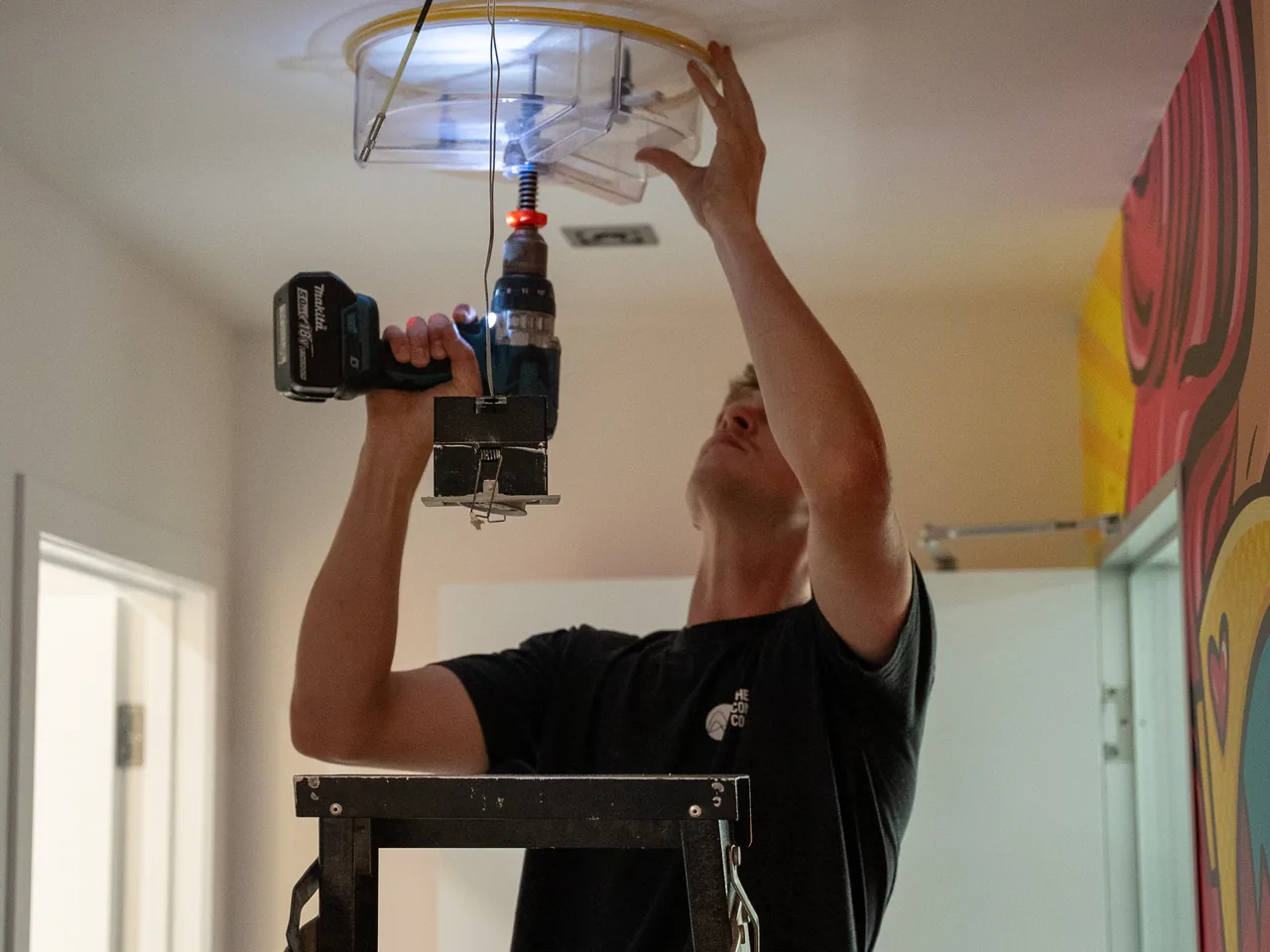 Electrician on a stepladder drilling a round speaker hole in a ceiling with a dust shroud