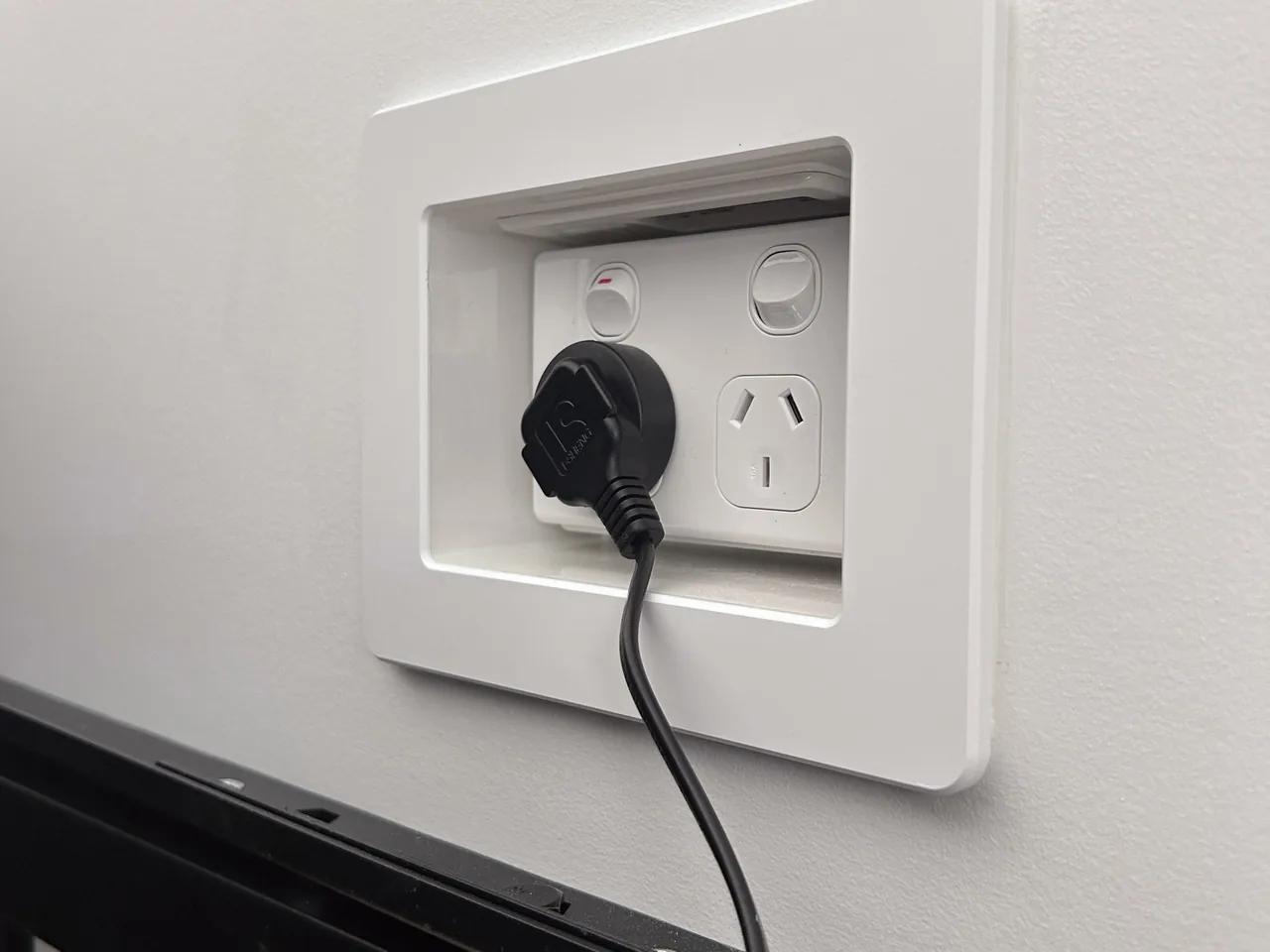Recessed power outlet set into a bedroom wall behind a TV, plug connected, tidy finish