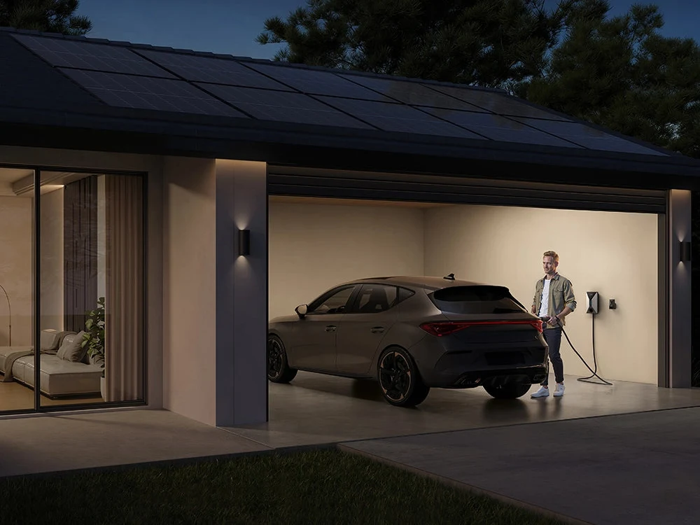 A man plugging his car into an Anker SOLIX V1 wall charger in a home garage at night, with solar panels on the roof