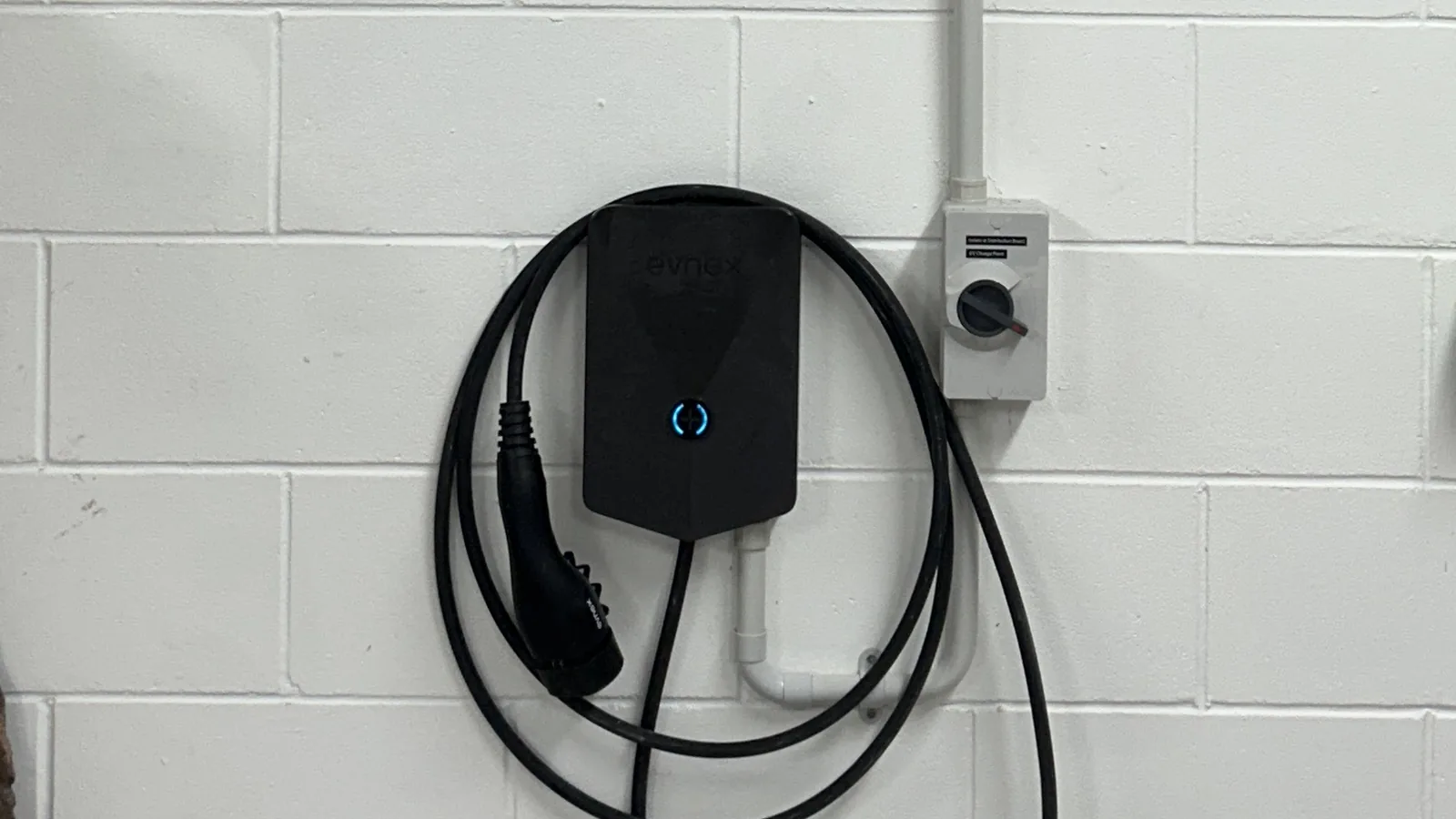 A wall-mounted EV charger with its charging lead coiled around it, beside an isolating switch on a garage block wall.