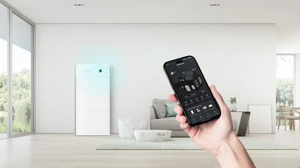 A white home battery cabinet against a living room wall, and a hand holding a phone showing its monitoring app.