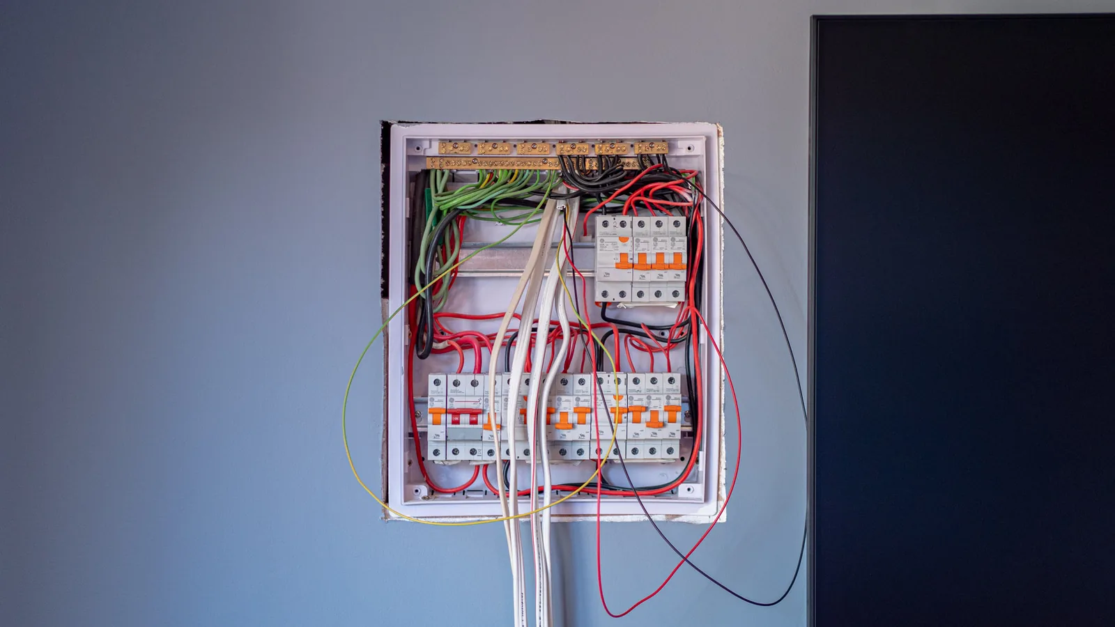 A recessed switchboard set into a cut wall opening mid-installation, with breakers fitted and cables still hanging loose