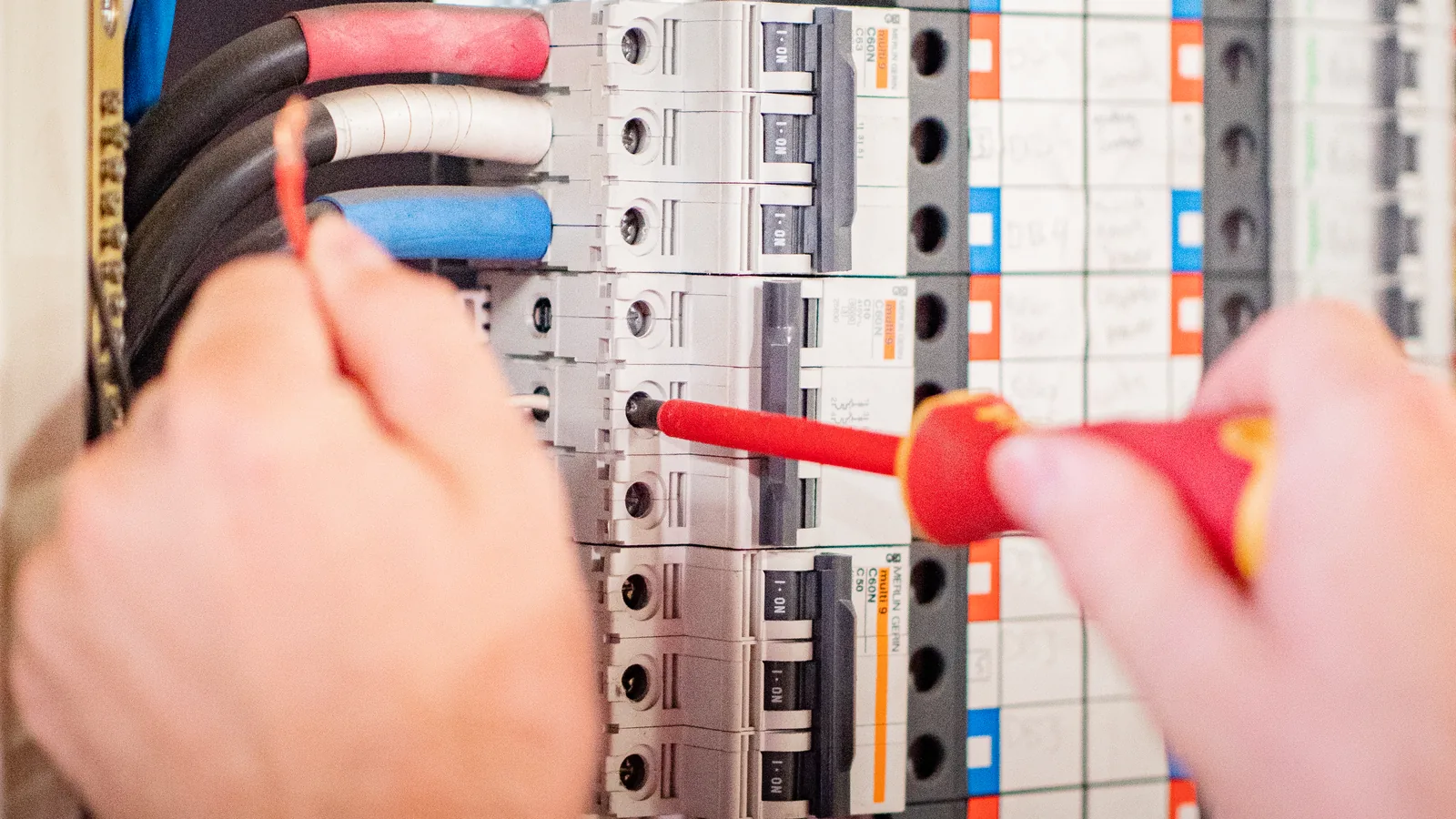 An insulated screwdriver tightening a terminal on a row of circuit breakers in a switchboard.