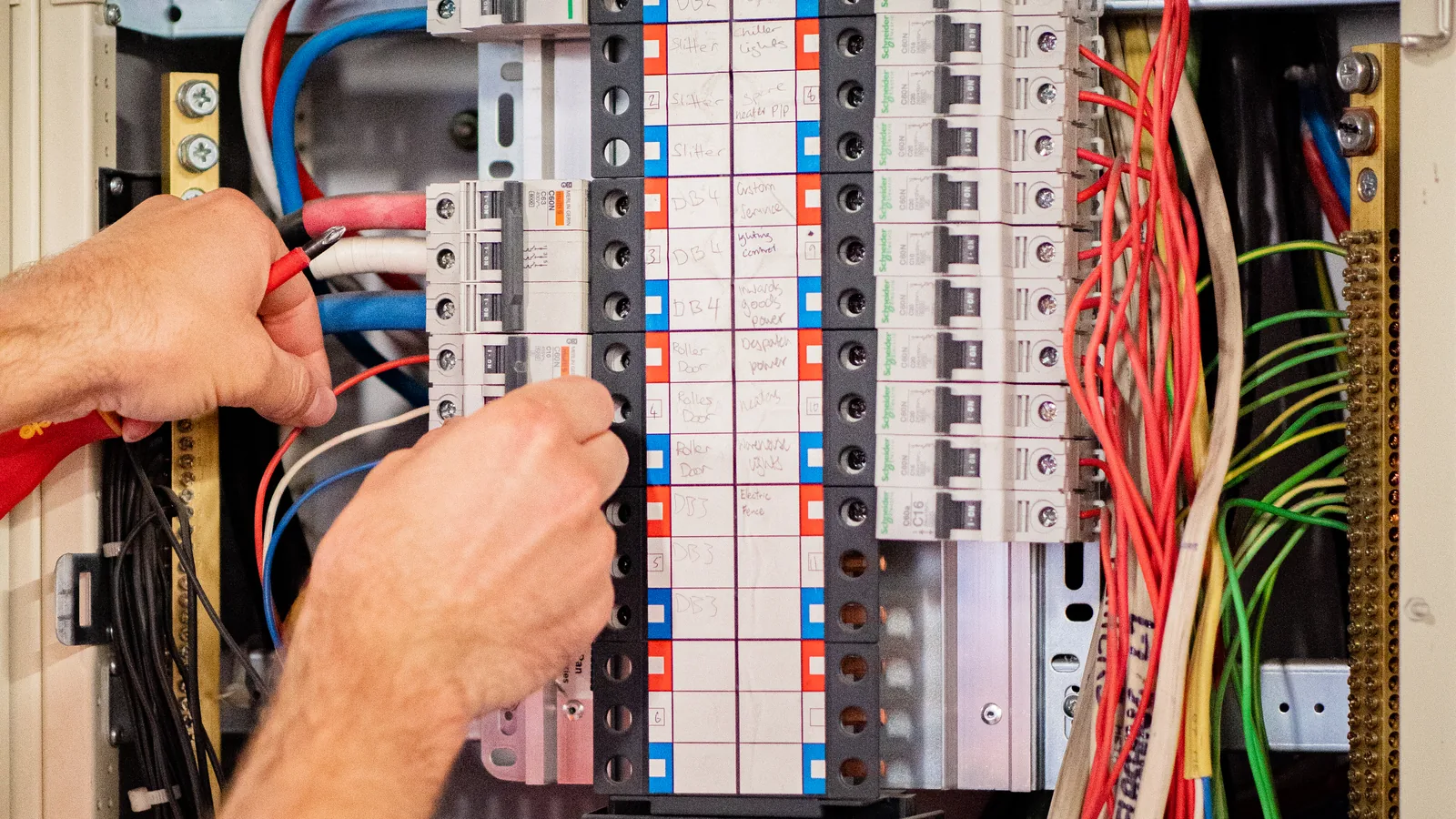 Hands lifting a cable tail inside an open switchboard fitted with two rows of circuit breakers.