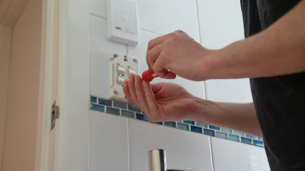 Hands with a red screwdriver fitting a switch plate to a tiled bathroom wall.