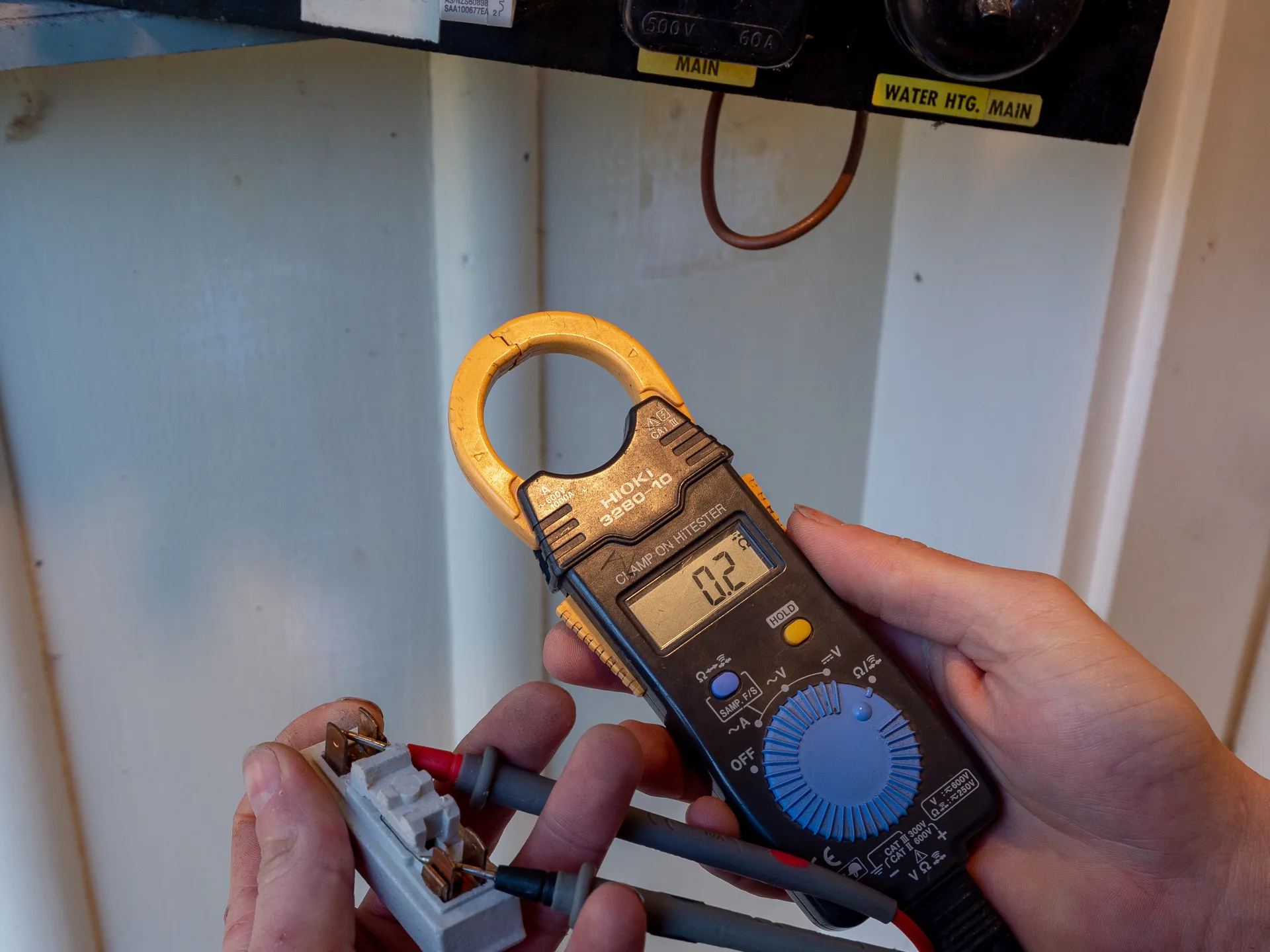 Hands holding a clamp meter and test leads under an old fuse board labelled water heating main