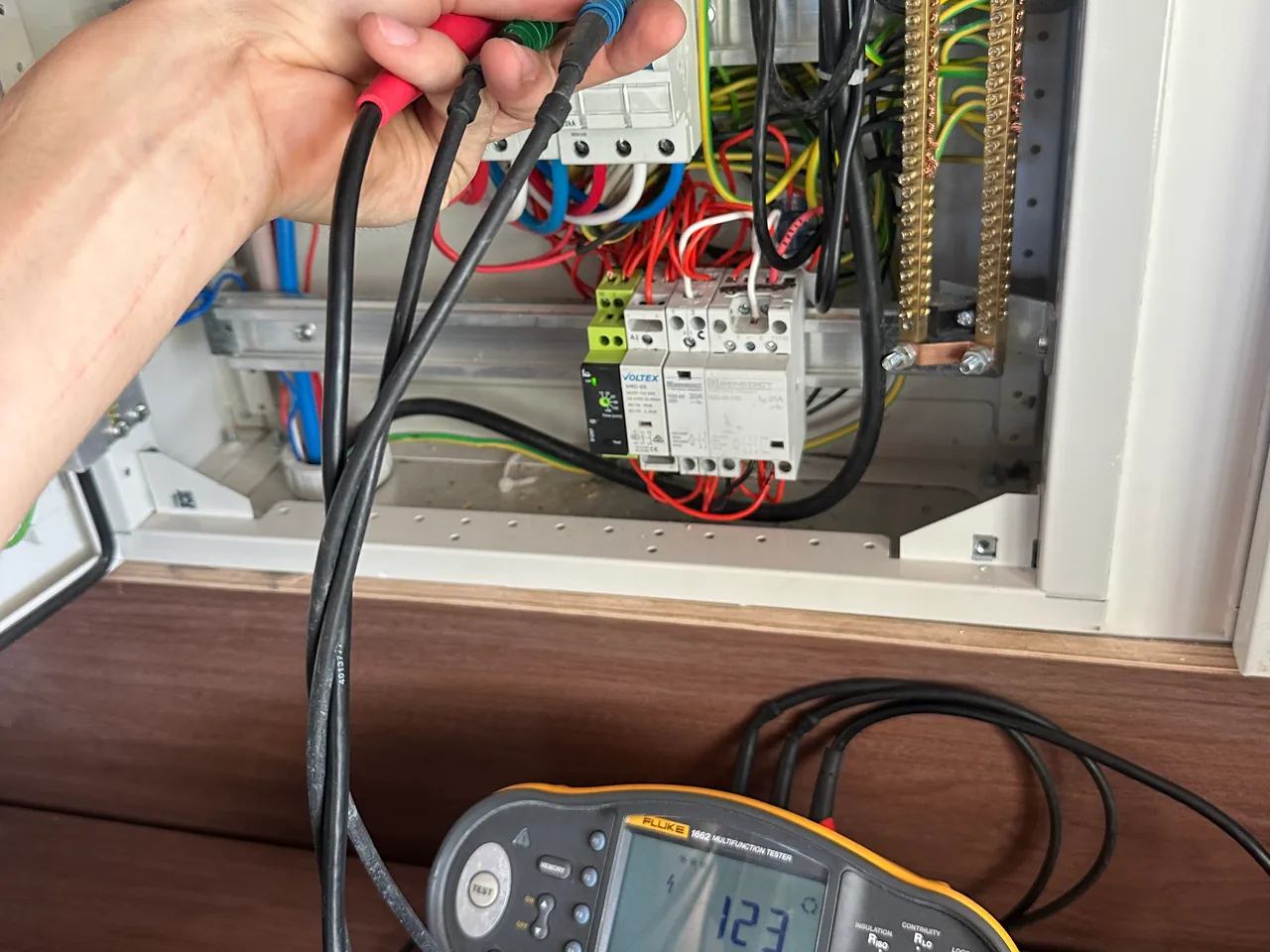 Hand holding red and black test probes against a distribution board, with a multifunction tester showing a reading