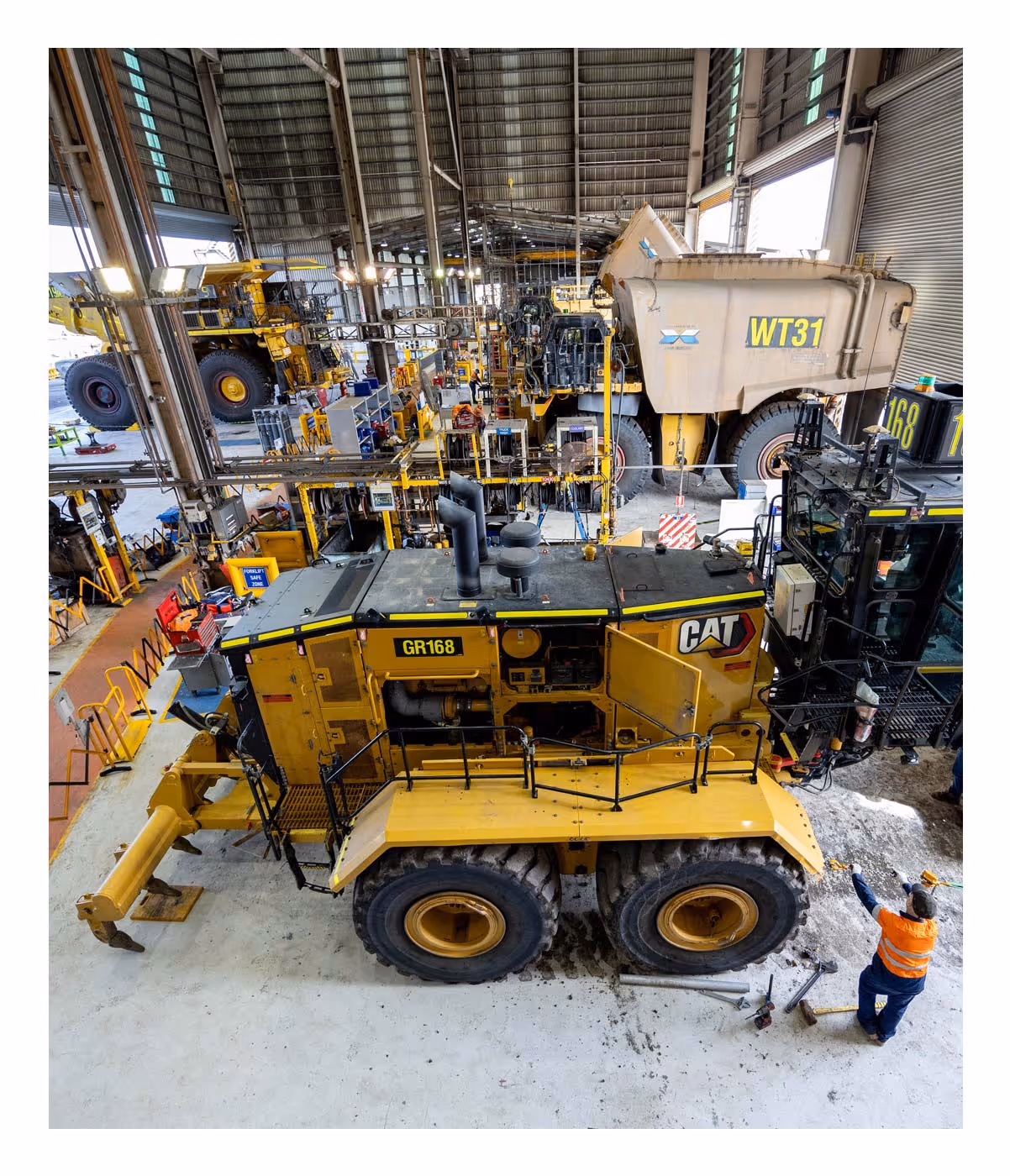 Large yellow CAT mining vehicle in a workshop with a worker in an orange vest nearby.