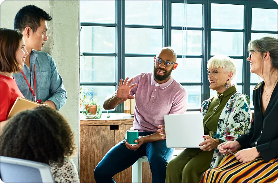 Diverse group of five colleagues discussing in a bright office with large windows, one man holding a coffee mug while gesturing.