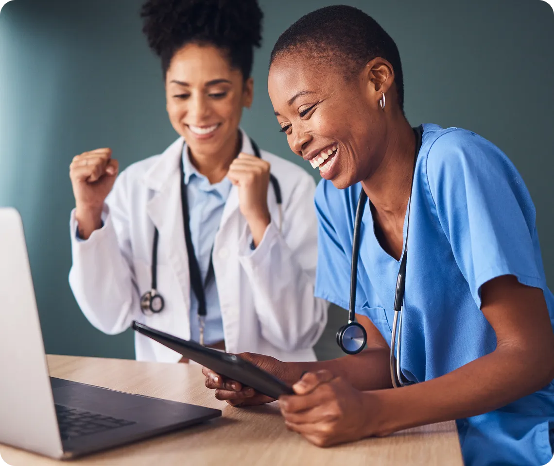 Two healthcare professionals smiling and celebrating, one in blue scrubs holding a tablet and the other wearing a white lab coat with a stethoscope around her neck, sitting at a table with a laptop.