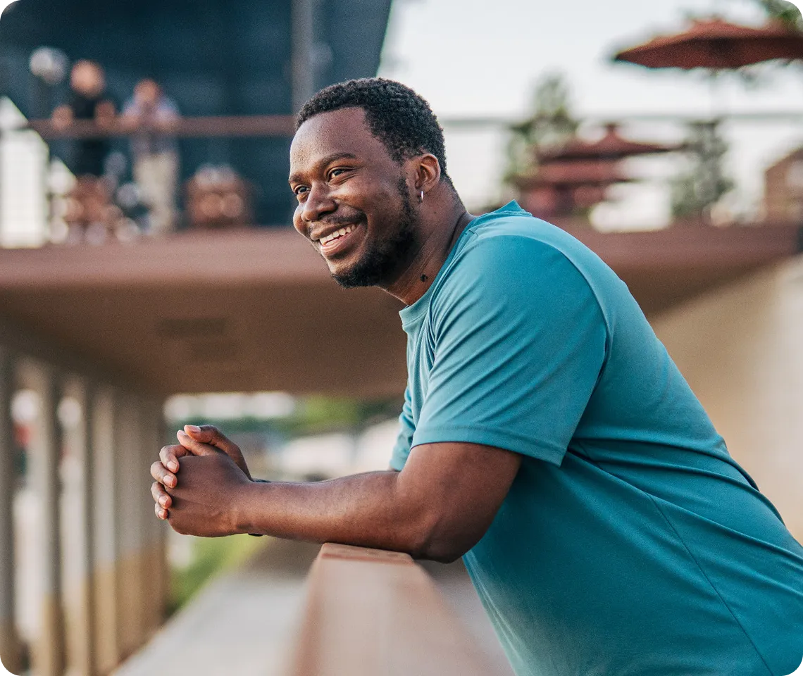 A smiling man in a teal shirt leaning on a wooden railing and looking to the side outdoors with blurred background featuring people and plants.