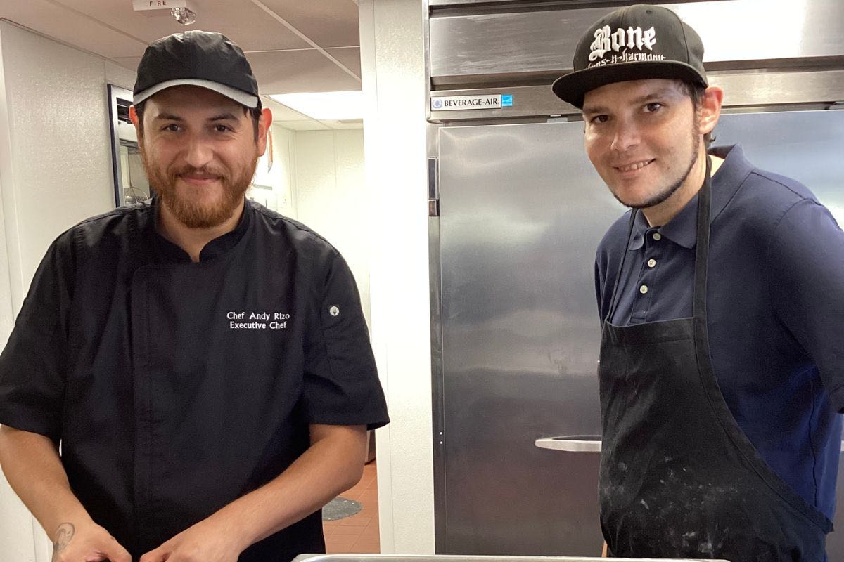 Two chefs in kitchen, one in black uniform and one in apron and hat