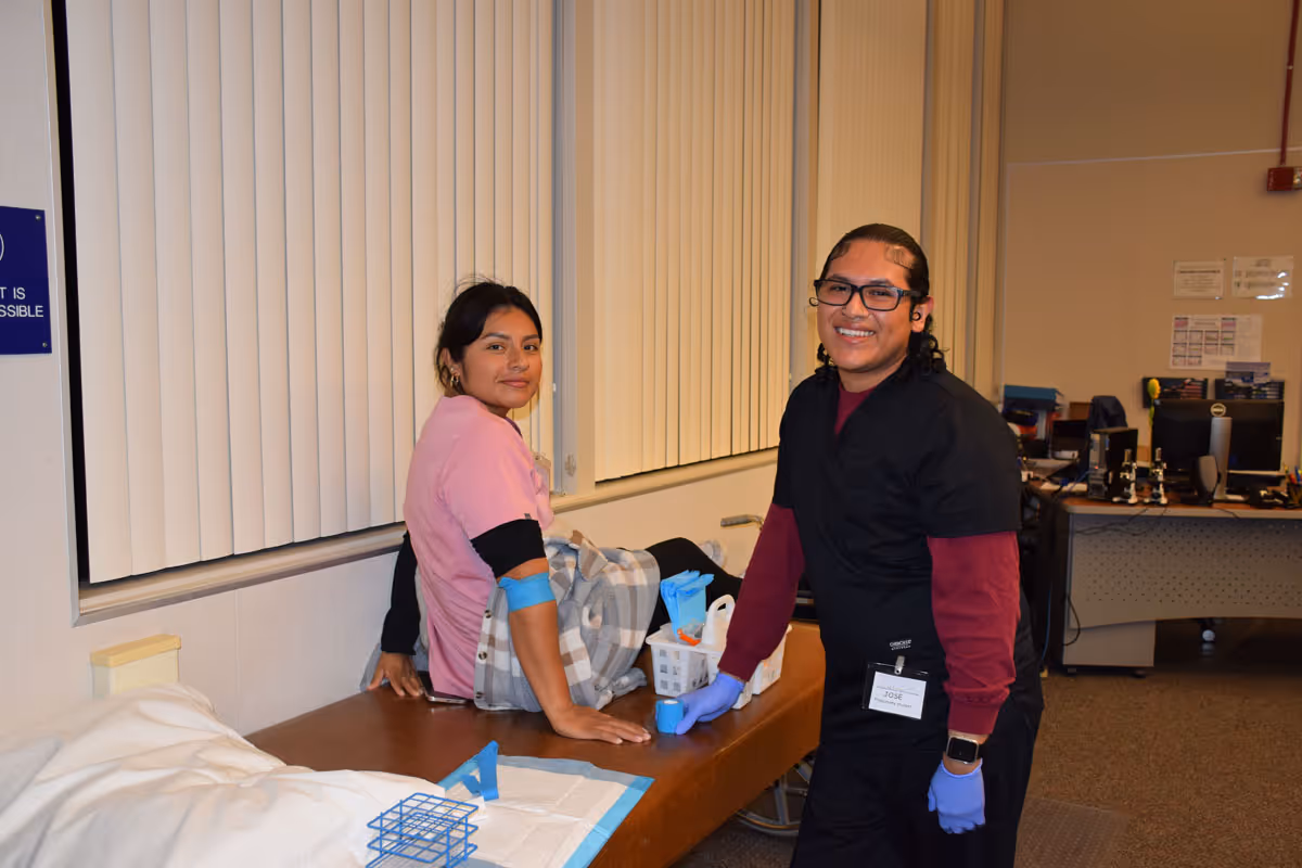 A woman in a pink medical uniform sitting on an examination table with a blue tourniquet on her arm, while a smiling healthcare worker in black scrubs and purple gloves attends to her in a clinical classroom setting.