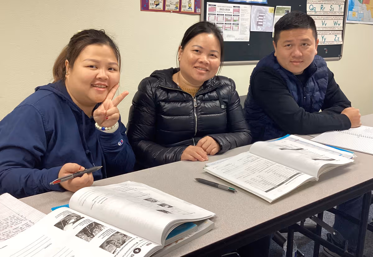 Three students sitting at a classroom table with open textbooks and notebooks, one woman making a peace sign.
