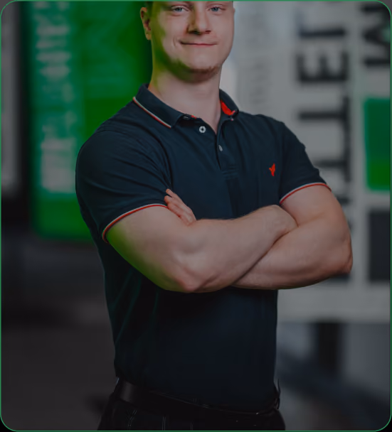 Portrait of a man with arms crossed wearing a black polo shirt with red trim, standing indoors with a green and black background.