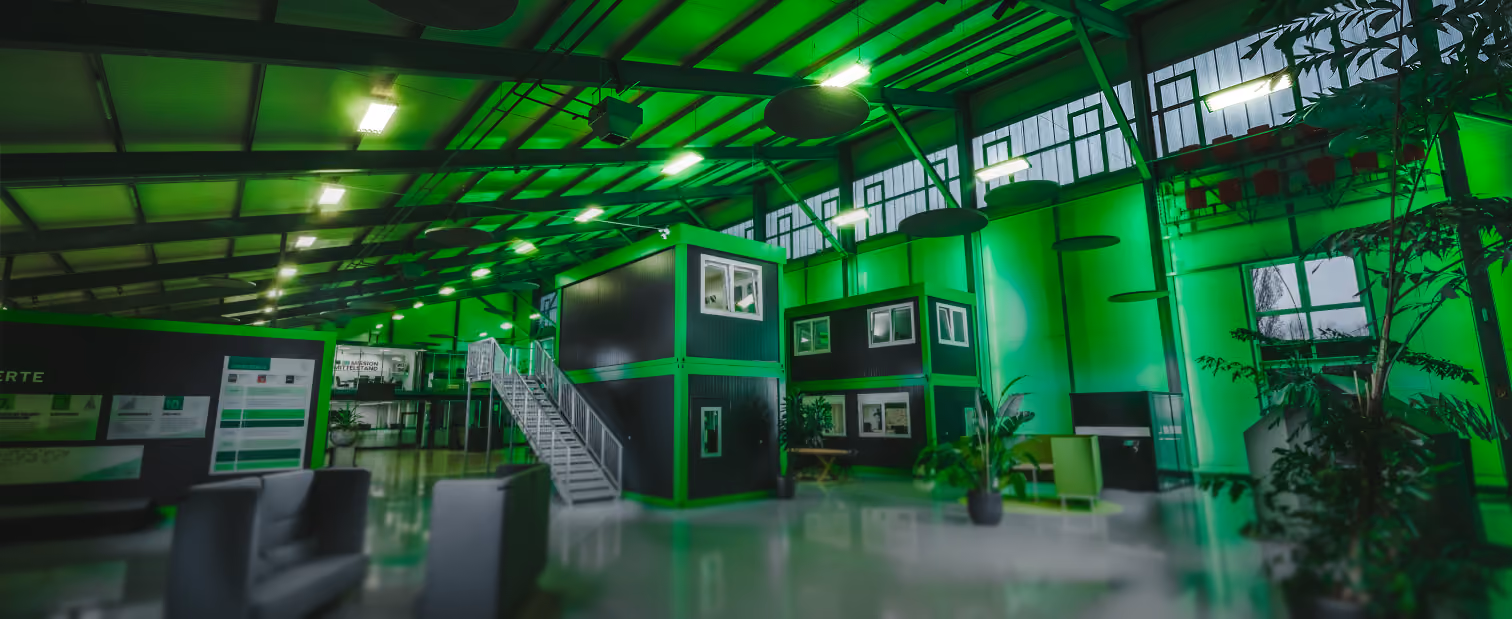 Interior of a modern industrial-style workspace featuring stacked modular office units with windows and an exterior staircase, illuminated by green lighting, with potted plants and seating areas inside a large warehouse with high ceilings and exposed beams.