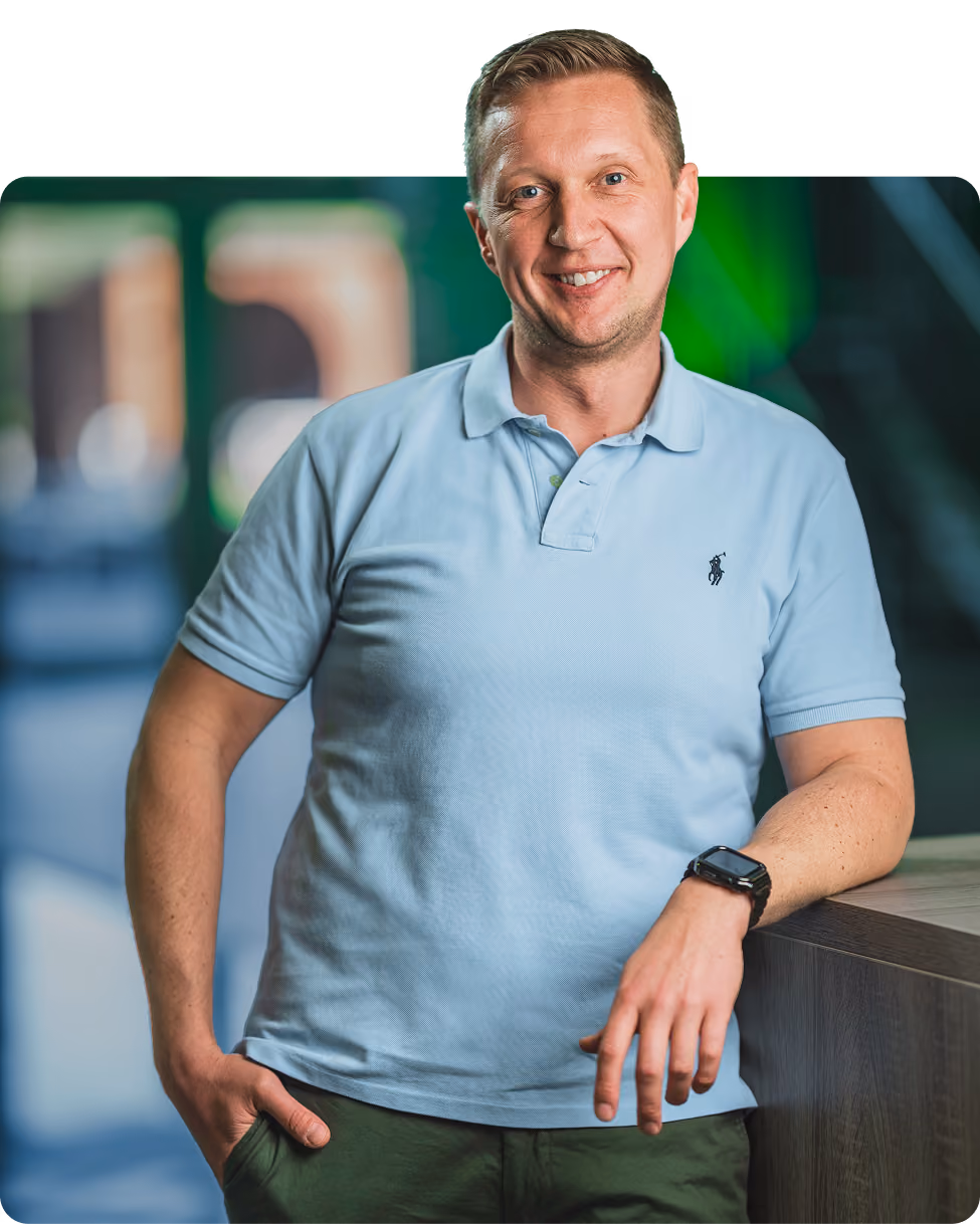 Smiling man wearing a light blue polo shirt and dark wristwatch, leaning on a wooden surface with one hand in pocket, indoors with blurred background.