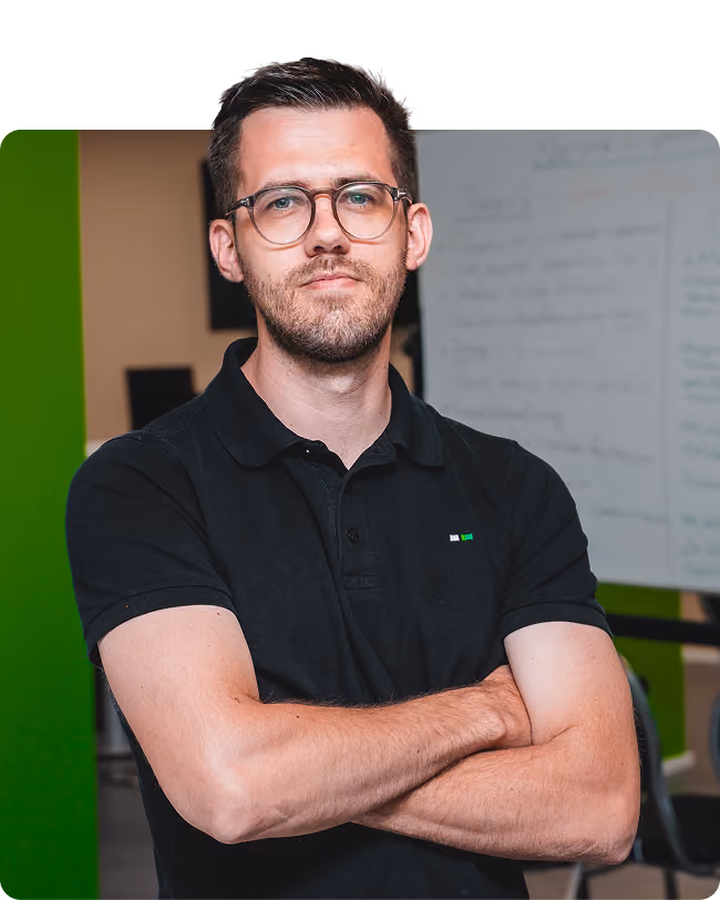 Man with short dark hair, glasses, and facial hair wearing a black polo shirt with arms crossed, standing indoors with a whiteboard and green wall in the background.