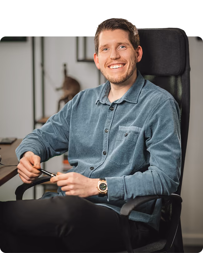 Smiling man with short brown hair and a beard sitting in a black office chair, wearing a blue button-up shirt and black pants, holding a pen in his hands.