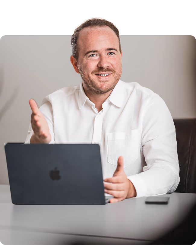 Man in a white shirt sitting at a desk with a laptop, gesturing with one hand while looking at the camera and smiling.