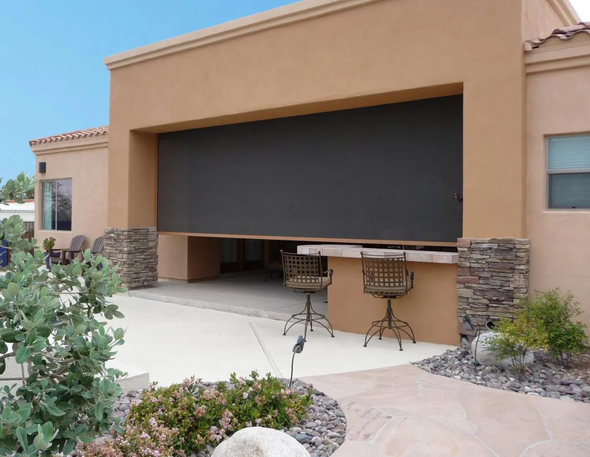 Outdoor patio with a large black retractable screen partially lowered, two bar stools at a counter, and surrounding landscaping with bushes and rocks.
