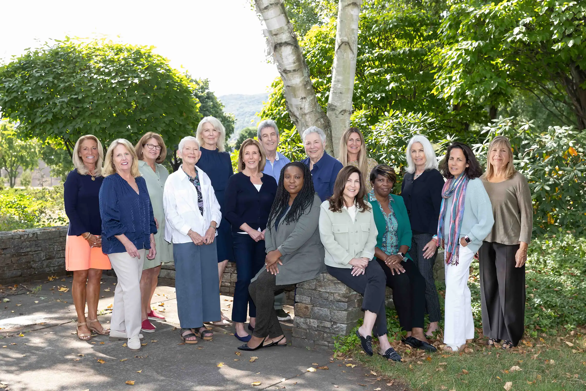 Sage Solutions employees pose outdoors in front of green trees and a stone wall, dressed in business casual attire, on a sunny day.
