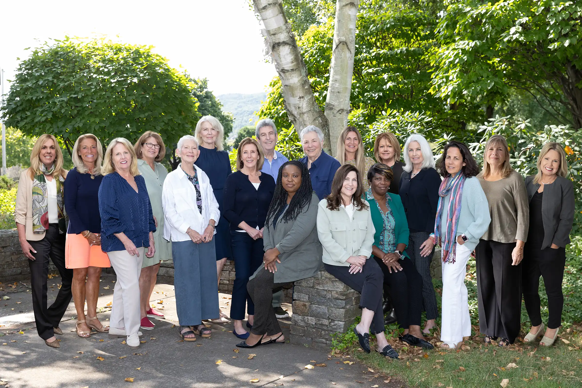 Sage Solutions employees pose outdoors in front of green trees and a stone wall, dressed in business casual attire, on a sunny day.