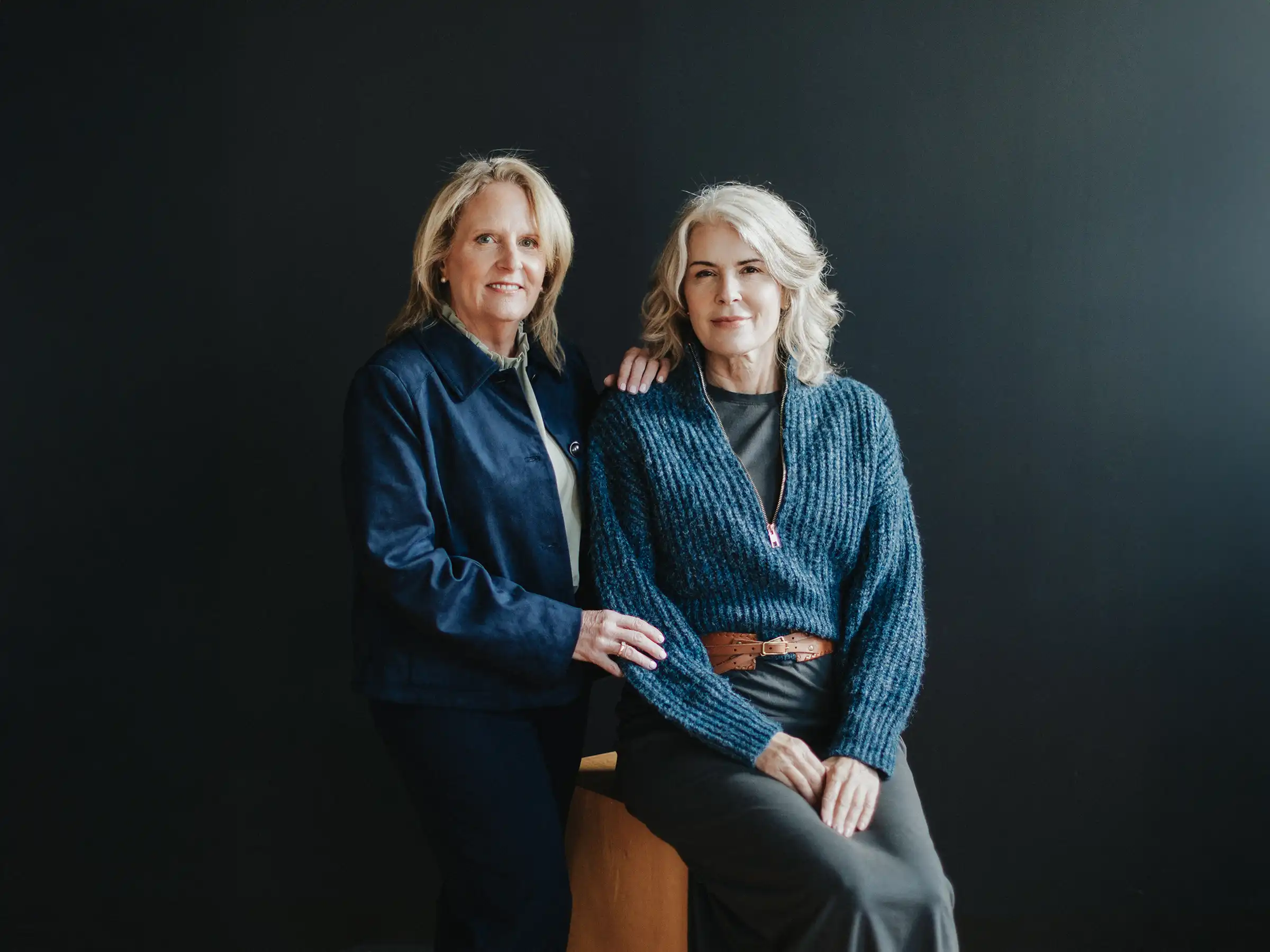 Valerie stands in a navy jacket with her hand on Tully's shoulder, while Tully sits wearing a blue knit sweater against a solid dark background.