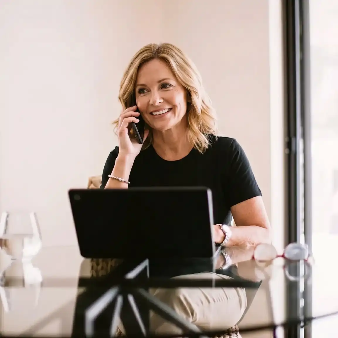 A blonde woman is seated at a glass table and on her smartphone. In front of her sits an open laptop, a glass of water, and eyeglasses
