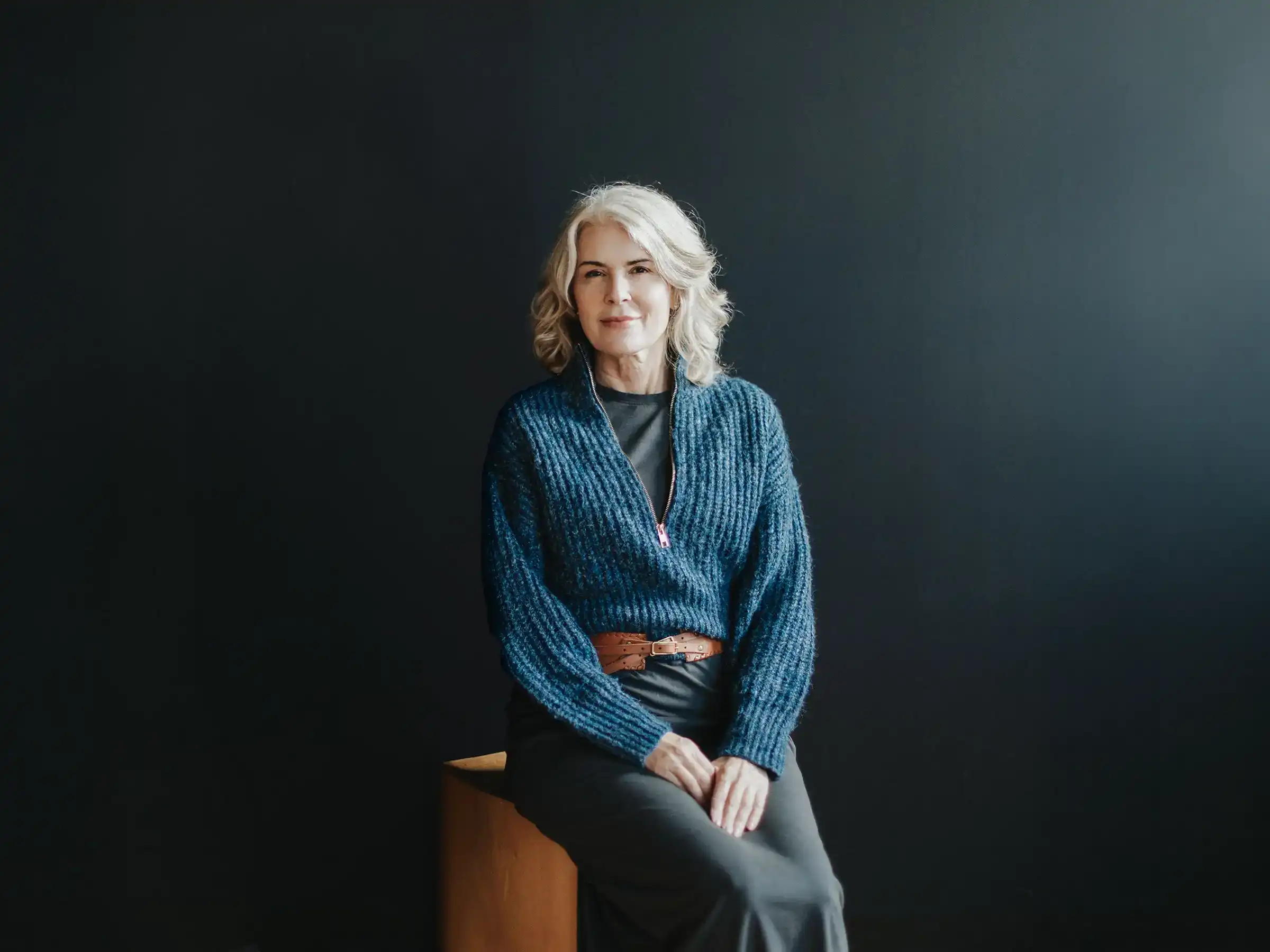 Tully Meyer sits on a wooden stool wearing a blue ribbed sweater and a brown leather belt against a dark, moody background.