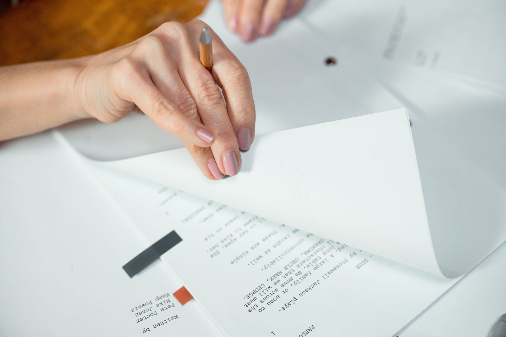 Person's hand with pale pink nails holding a pencil and turning pages of printed documents.