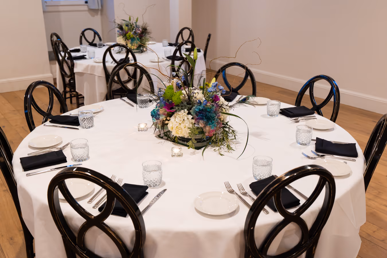 Round tables with white tablecloths set for an event, each with a floral centerpiece, glassware, black napkins, and silver cutlery.