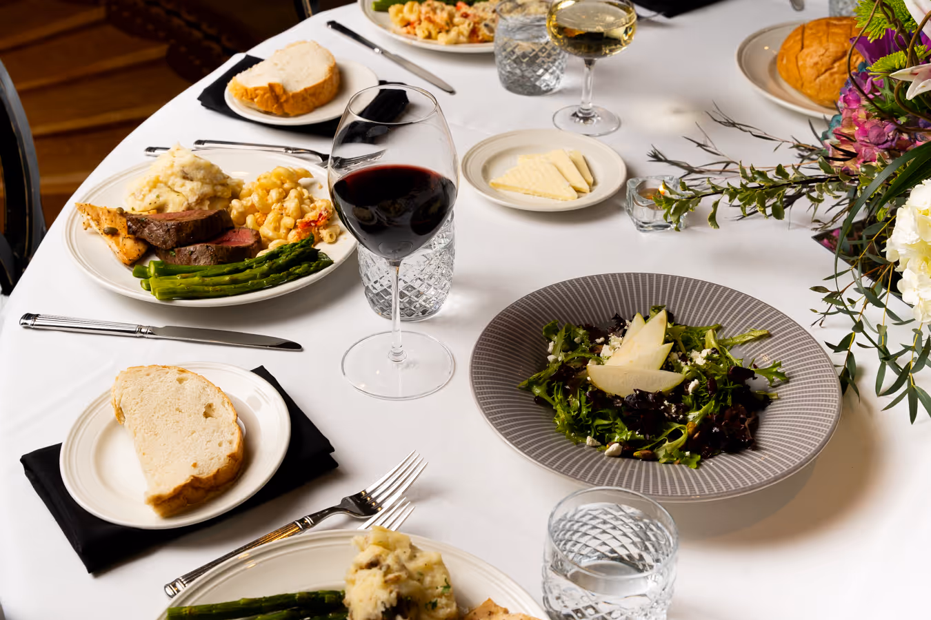 Elegant dining table with plates of steak, asparagus, pasta, salad with pear slices, bread, and glasses of red and white wine.