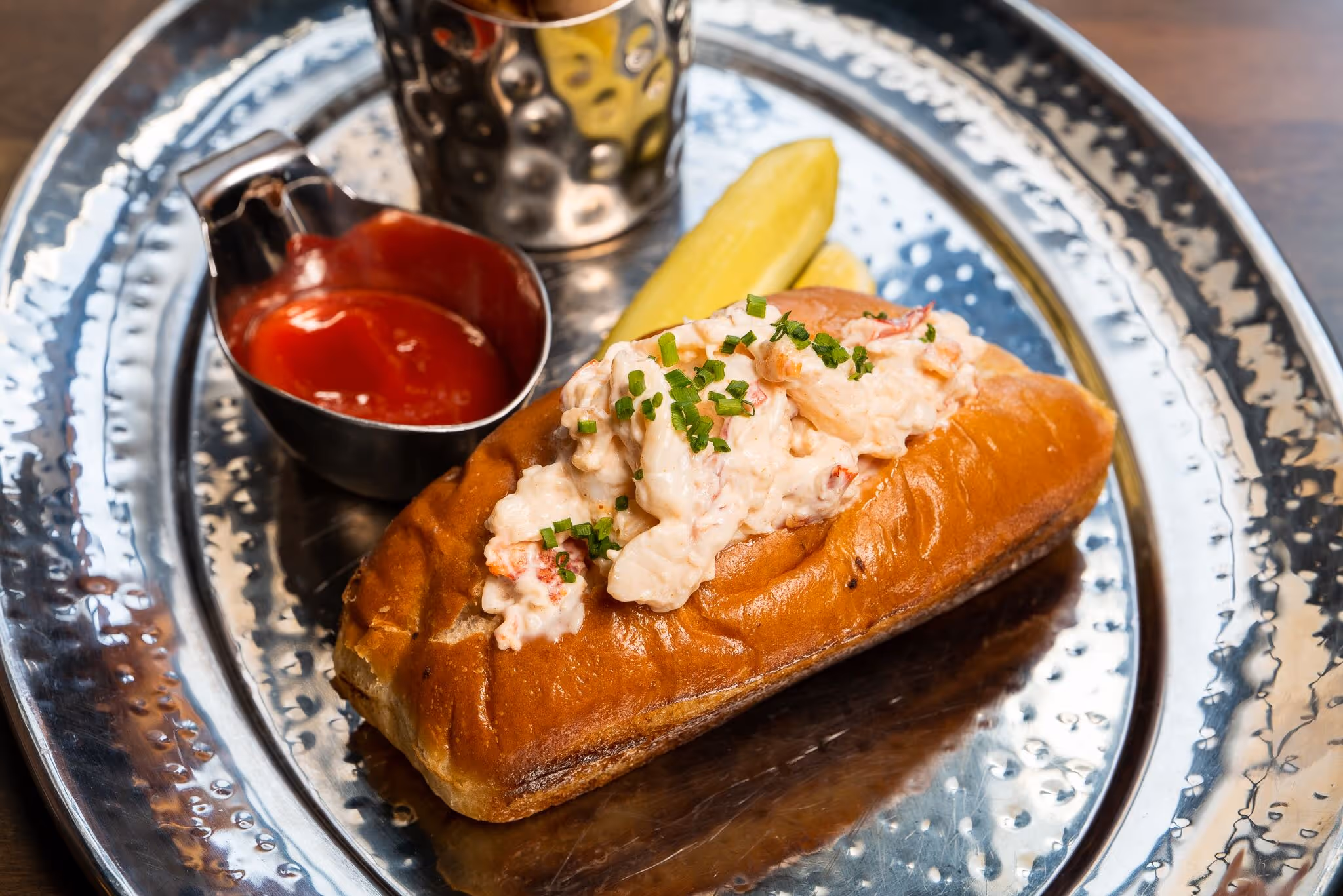 Lobster roll with creamy lobster salad topped with chopped chives, served with a pickle and a small cup of ketchup on a hammered metal plate.