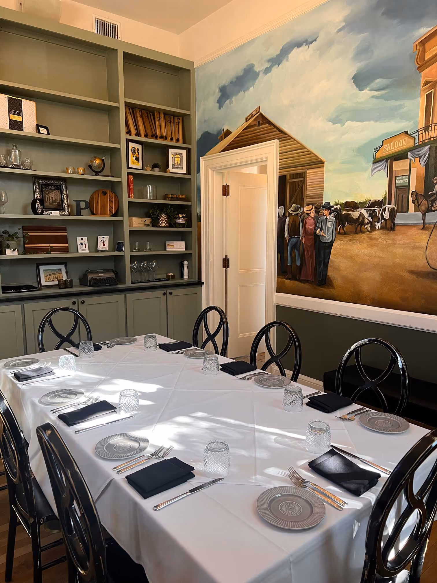 Dining room with a white tablecloth-covered table set with plates, silverware, folded black napkins, and glasses, surrounded by black chairs, with a mural depicting a western town scene on one wall and built-in shelves on another.