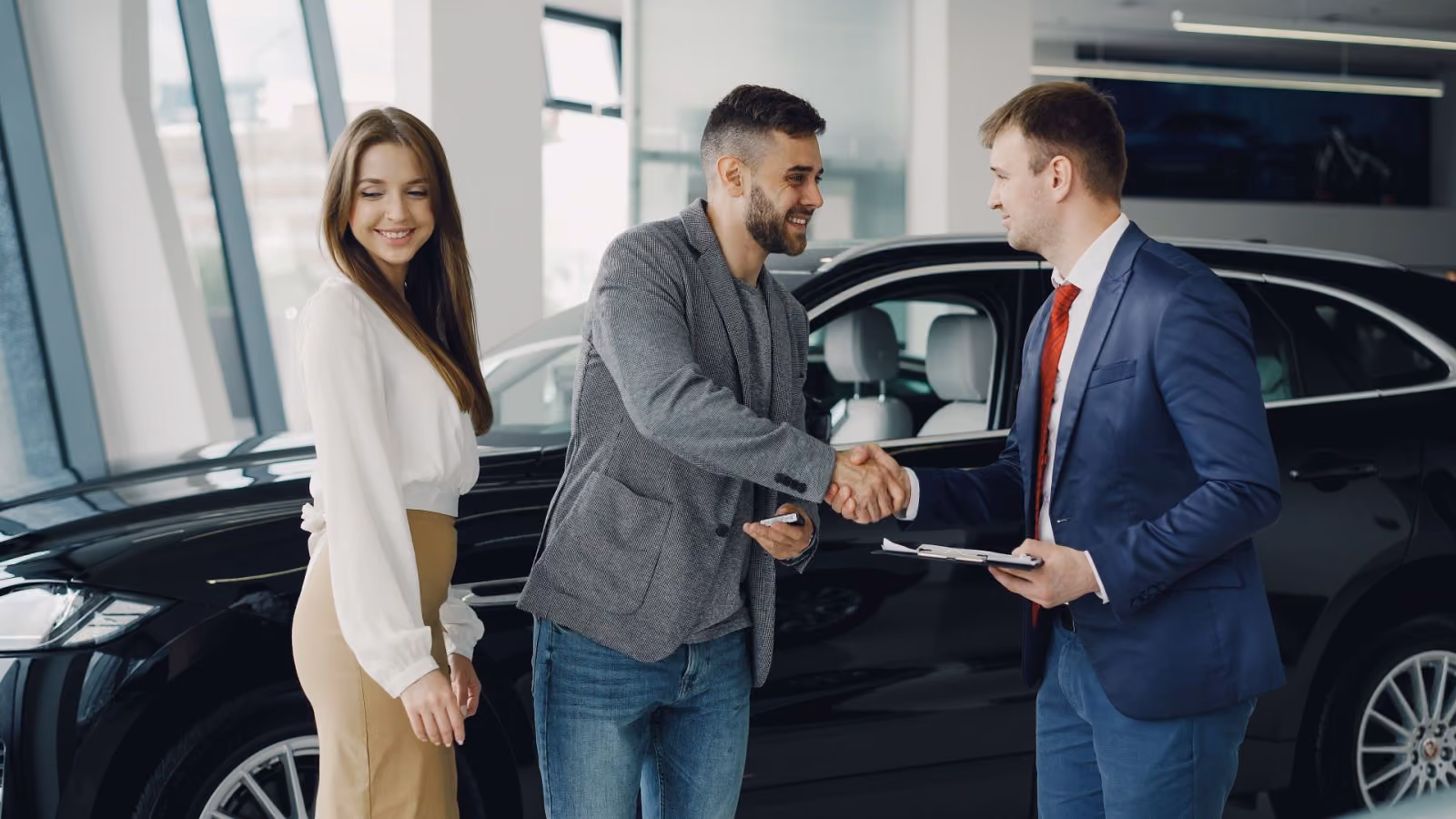 Man shaking hands with a car salesman, woman standing next to a black car in a showroom.