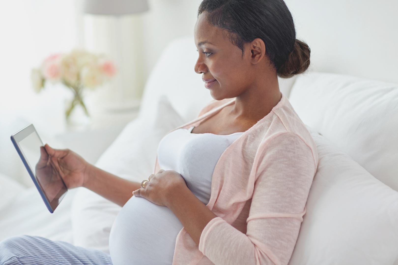 Black pregnant person in a white tank top and pink cardigan reading a tablet in bed and resting her hand on her growing belly