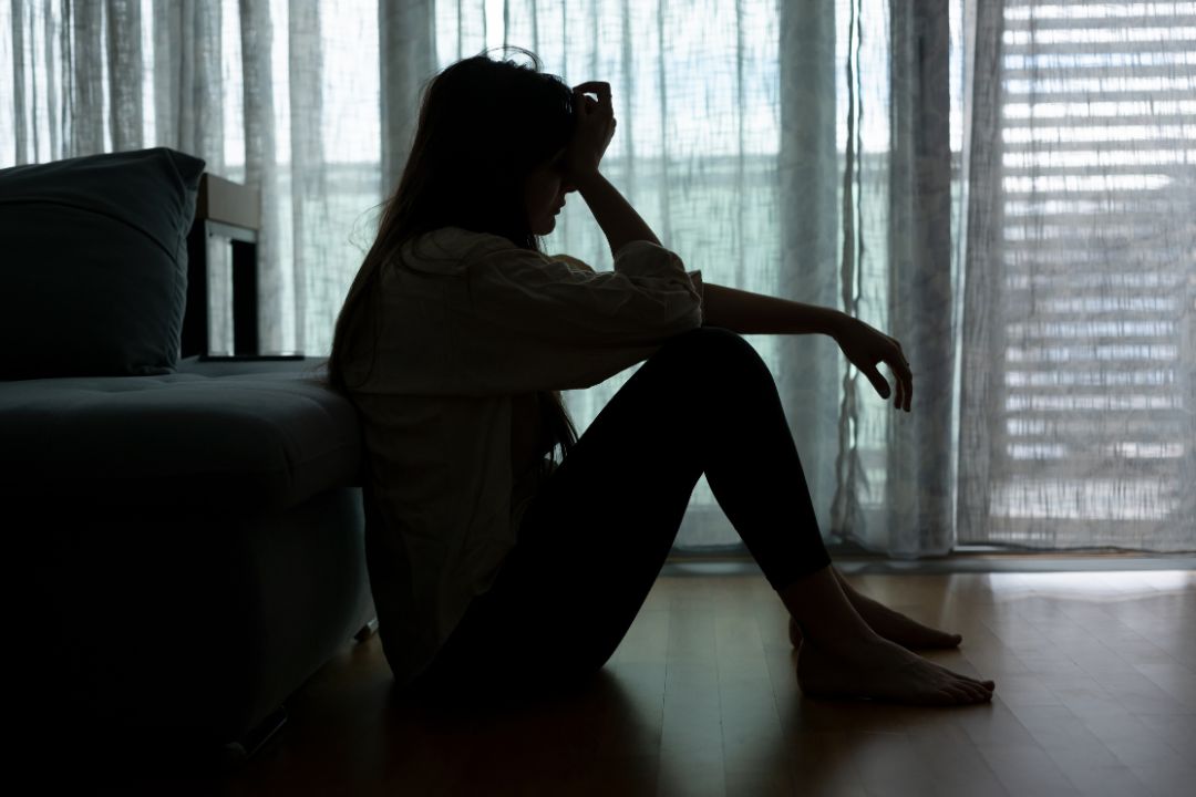 Woman sitting on the floor with her back against a bed and holding her head in her hands