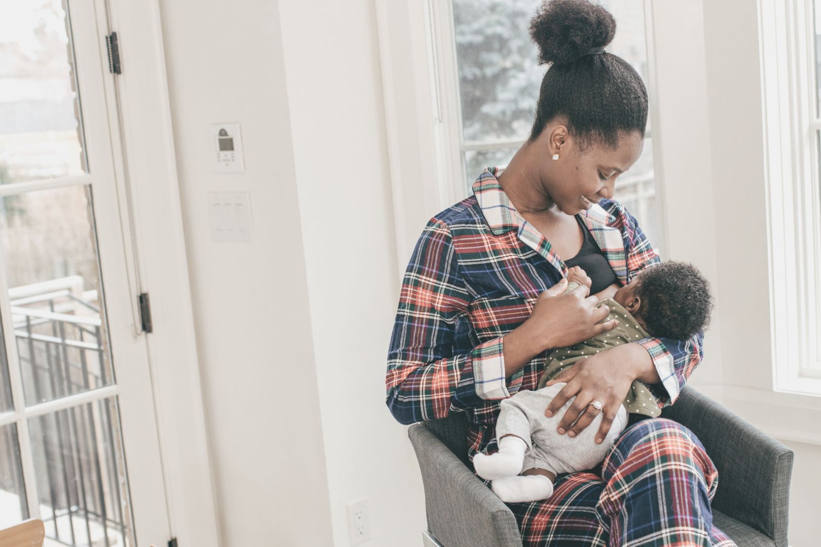 black mother nursing infant sitting in apartment by windows wearing blue plaid pajamas