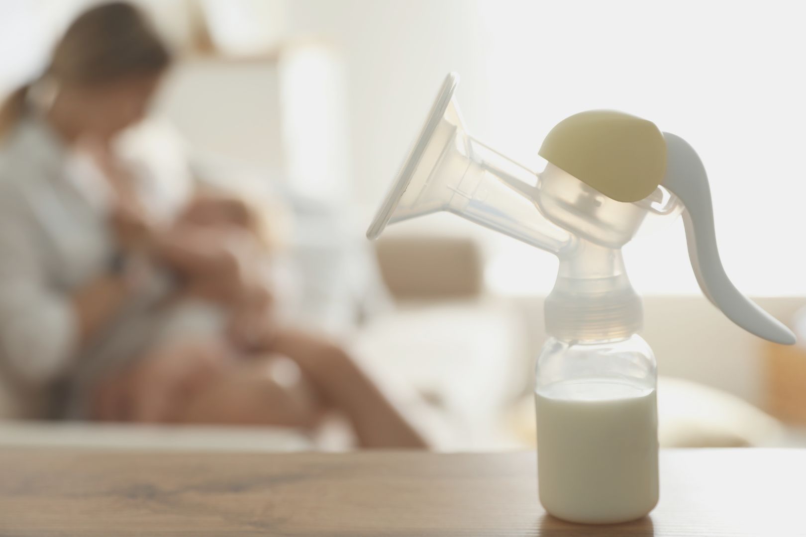 manual breast pump bottle full of milk with a mother nursing infant in the background
