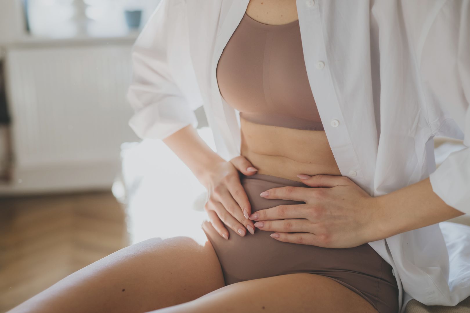 Woman holding her abdomen in taupe underwear and an open button up shirt