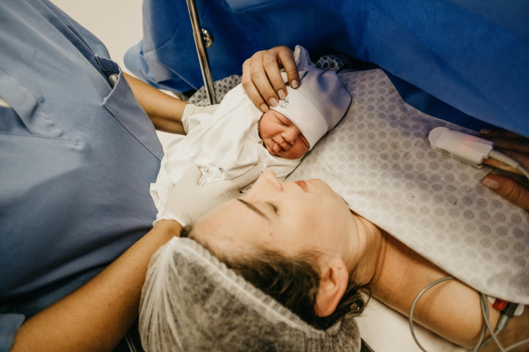 A woman in a hospital bed wearing medical gown and cap is handed her newborn from a healthcare worker