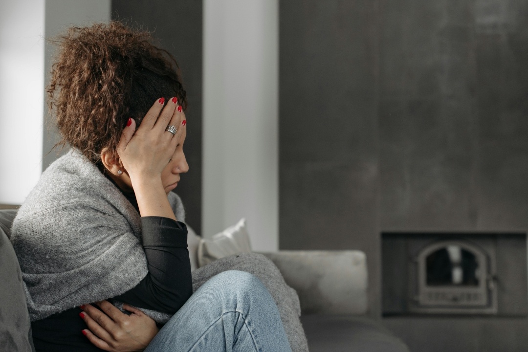 Worried postpartum woman sitting in a grey room, one hand on her temple and one on her belly