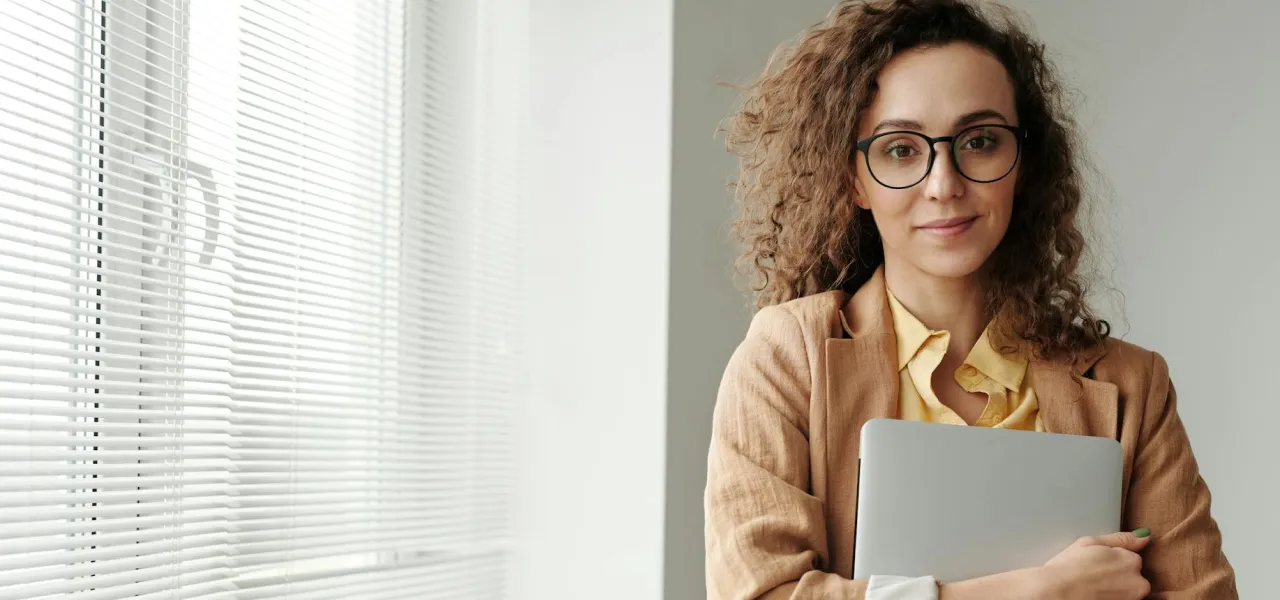 Woman holding closed laptop standing beside a window