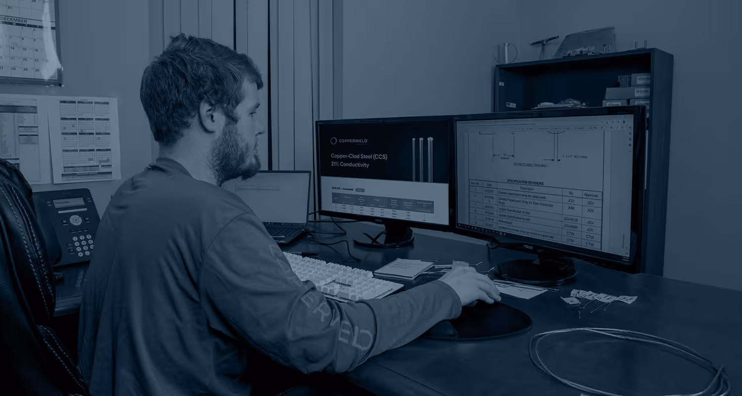 Man with beard works on dual monitors showing Copperweld data and technical specification revision sheet.